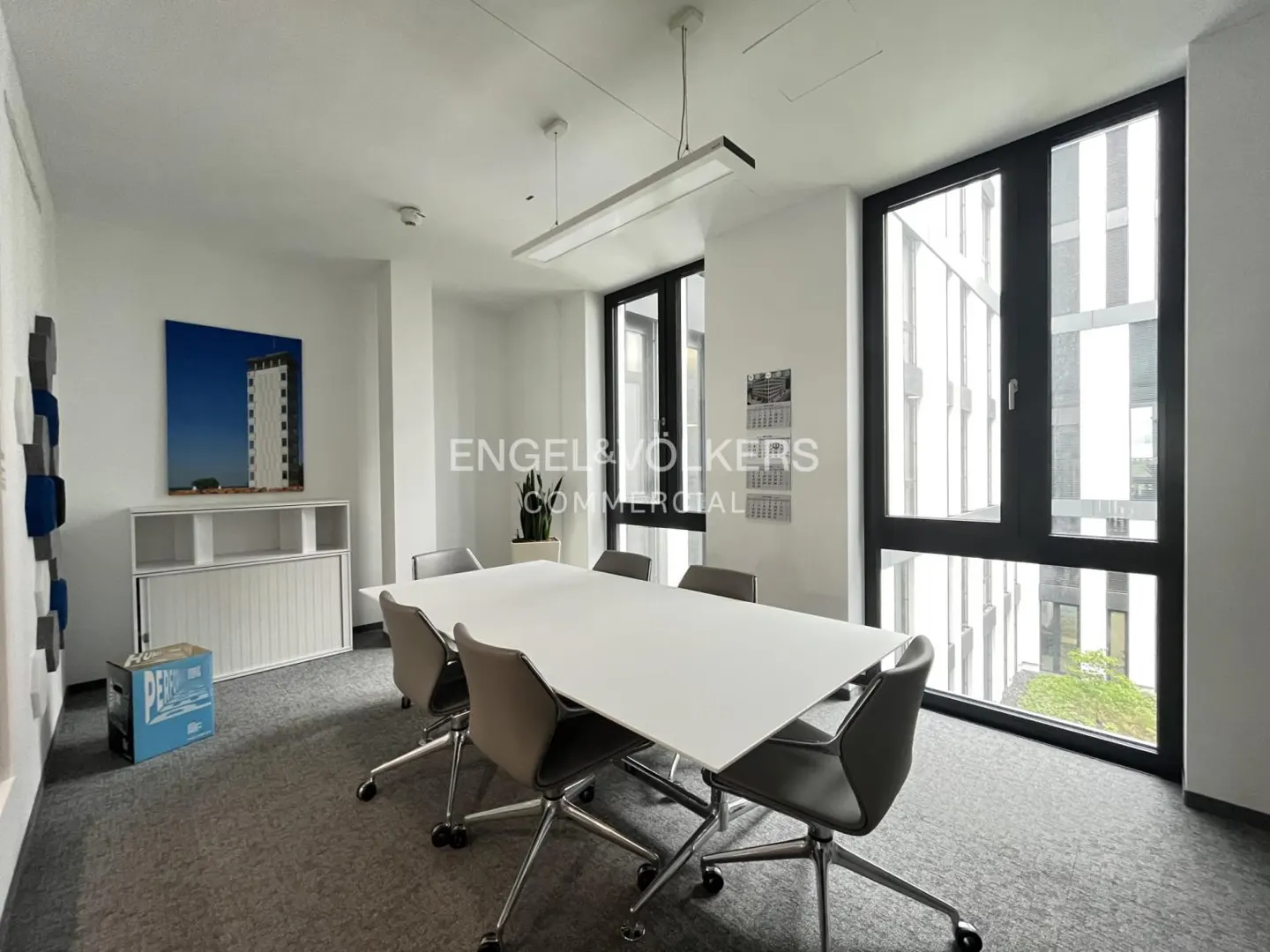 Bright office space with a white conference table, gray chairs, and large windows. A modern art piece and storage unit are visible.