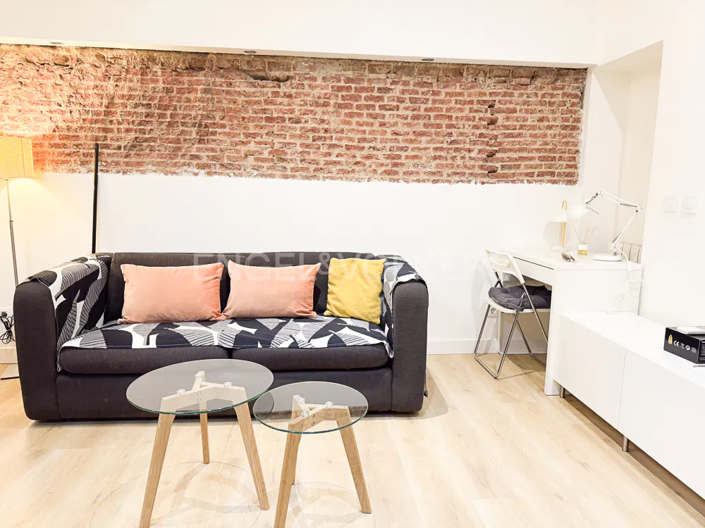 Living room with exposed brick wall, gray sofa with peach pillows, and glass coffee tables on a light wood floor.