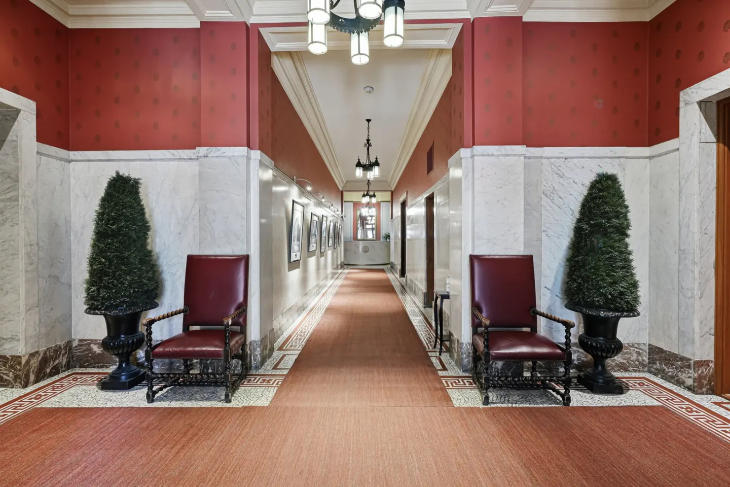 Long hallway with red carpet, marble walls, and red accent walls. Two red chairs flank the entrance, each with a topiary. Chandeliers hang from the ceiling.