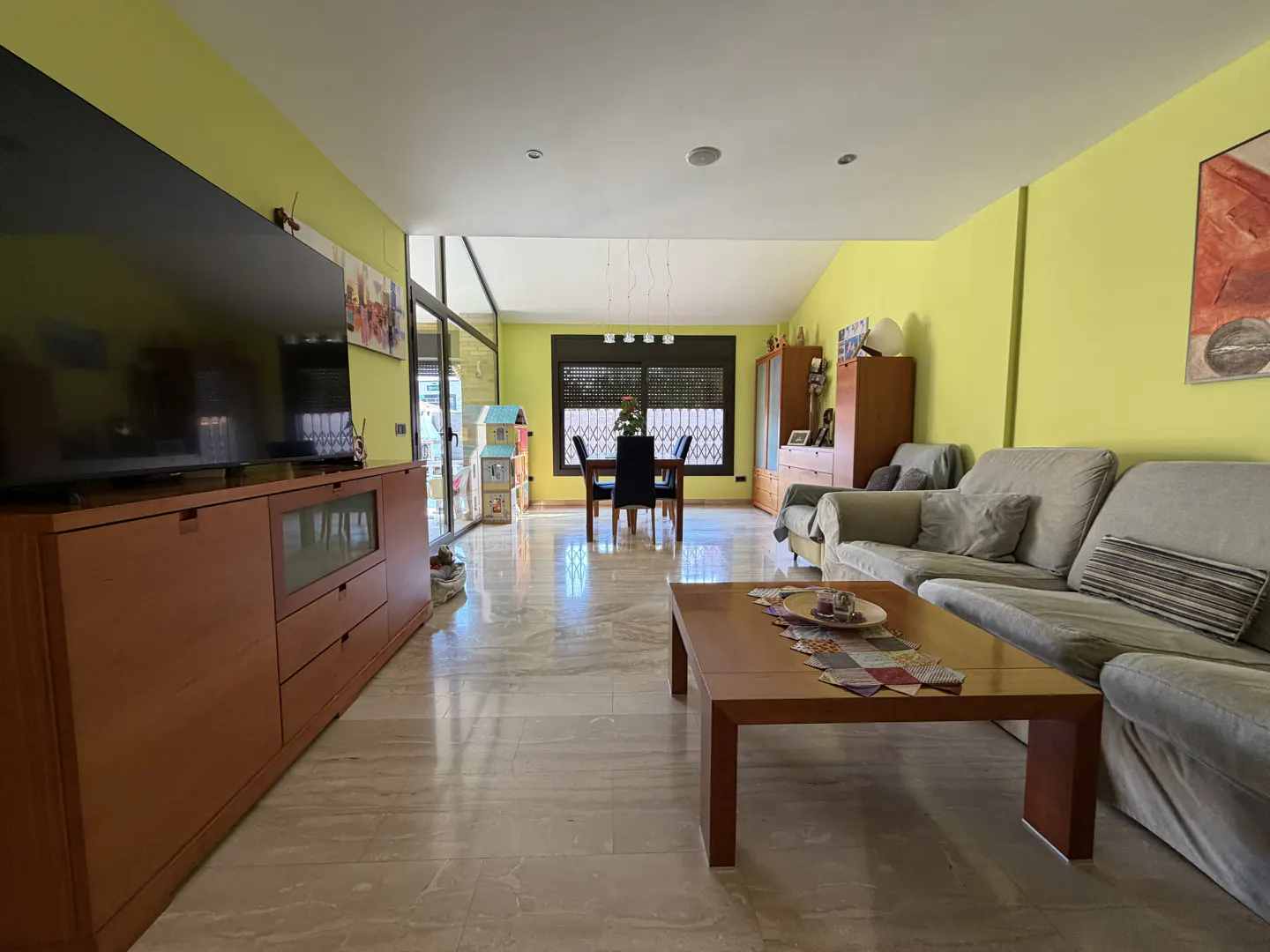 A bright living room with yellow walls, a large TV, a dining table, and a gray sofa. The floor is tiled with marble.