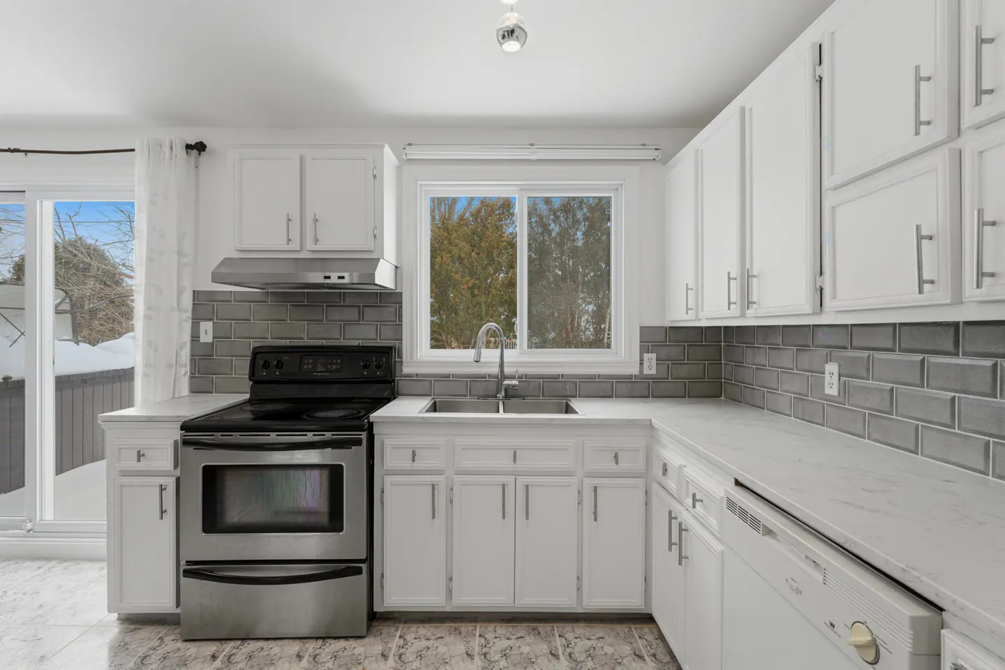 Bright kitchen with white cabinets, gray backsplash, and stainless steel appliances. A window overlooks a snowy outdoor scene.