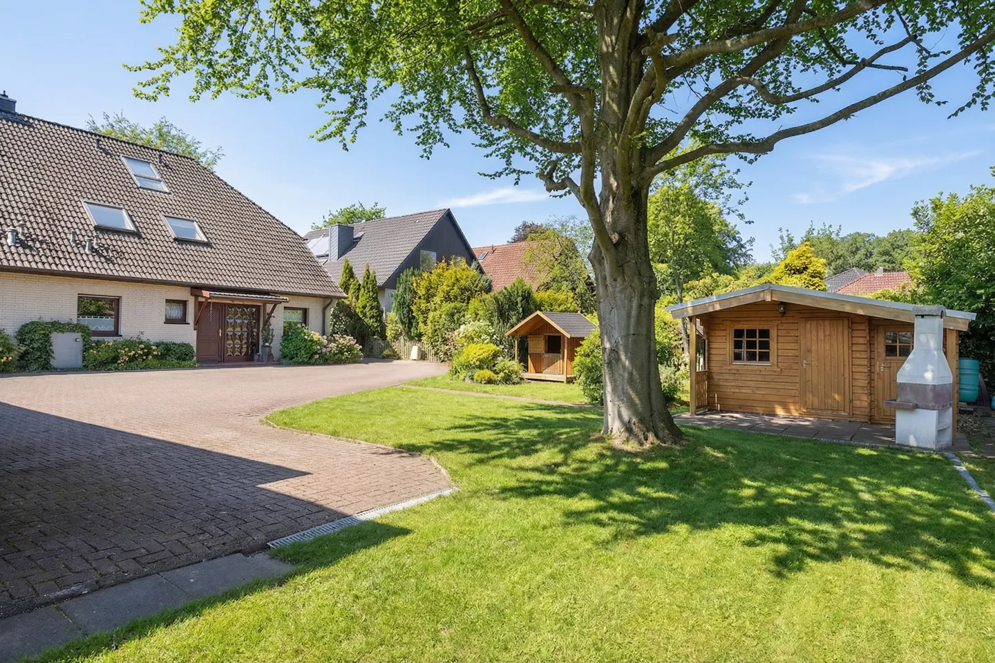 Exterior view of a house with a brick driveway, green lawn, a large tree, and two wooden sheds on a sunny day.