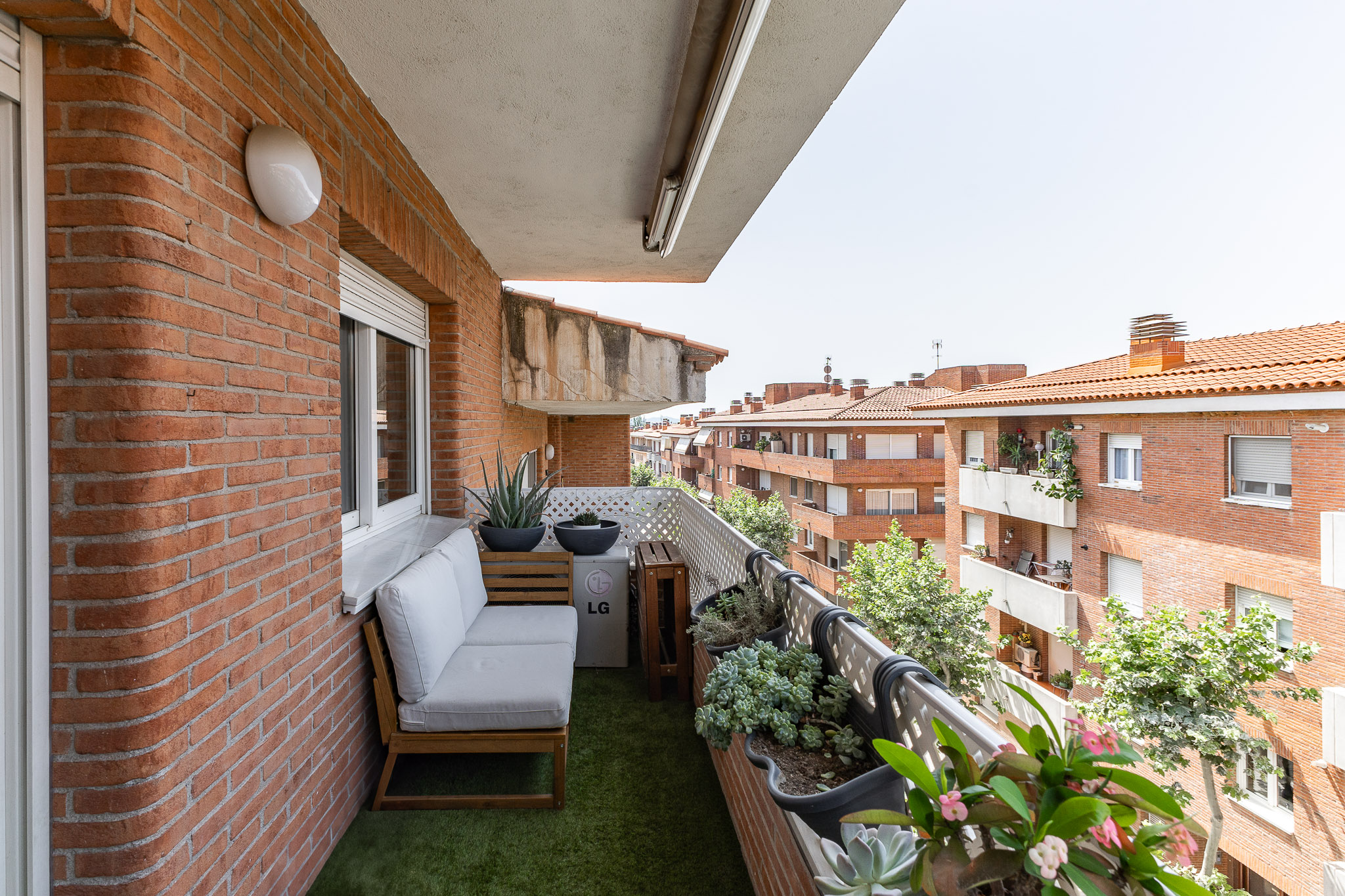 A balcony with a white sofa, artificial grass, potted plants, and a view of surrounding brick apartment buildings.