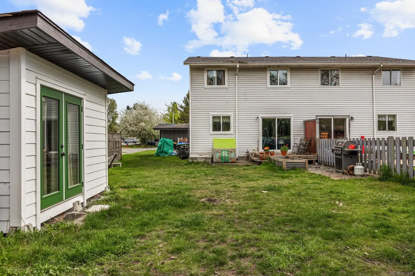 Backyard view of a two-story house with white siding, a shed with green doors, and a green lawn under a blue sky.