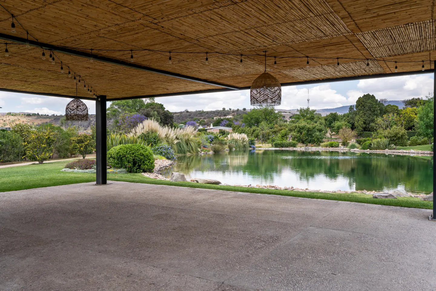 Covered patio with a bamboo ceiling, string lights, and wicker pendant lights overlooking a pond and lush green landscape.