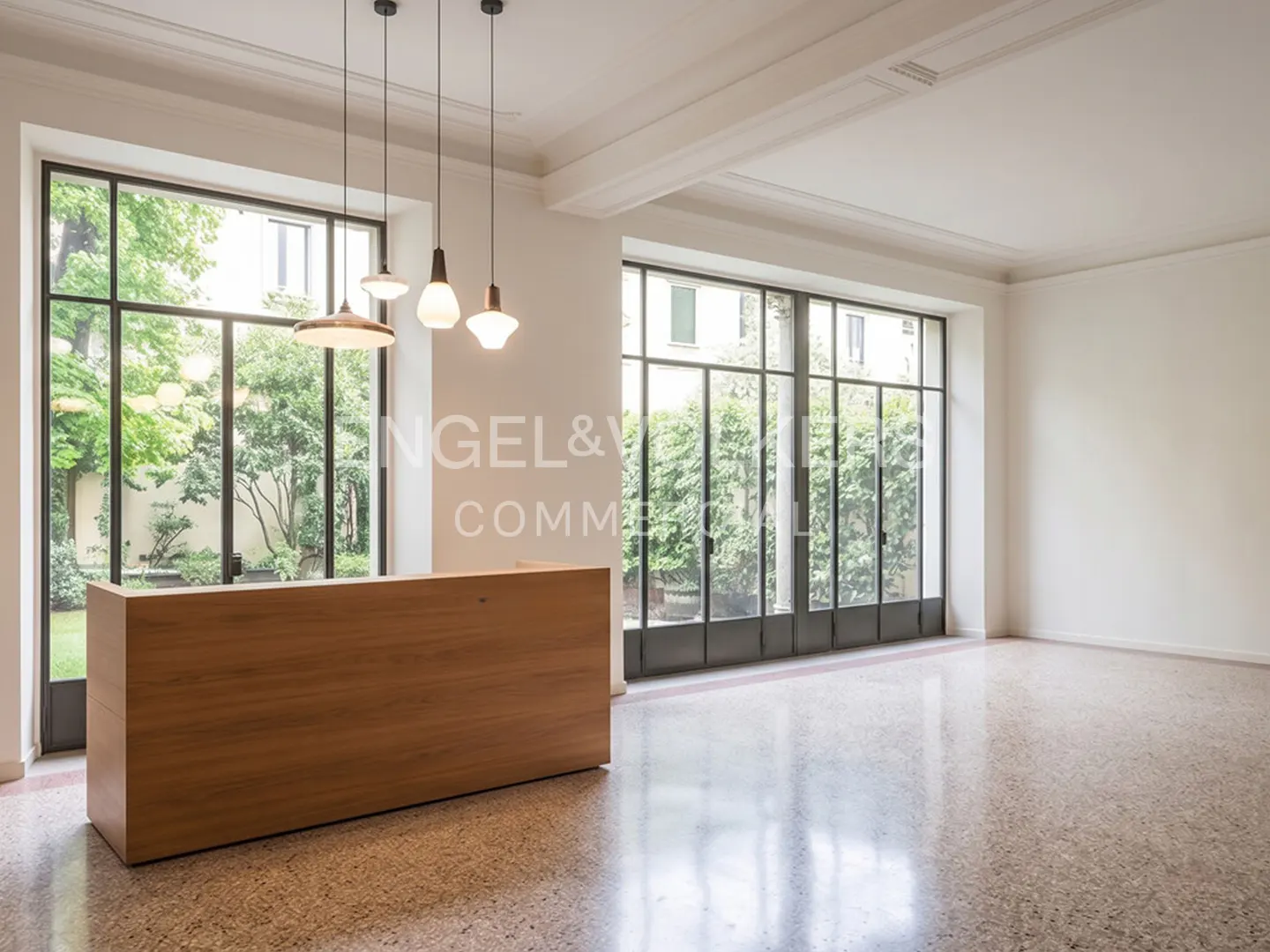 A bright, empty office space with a wood reception desk, pendant lights, and large windows overlooking a green garden.