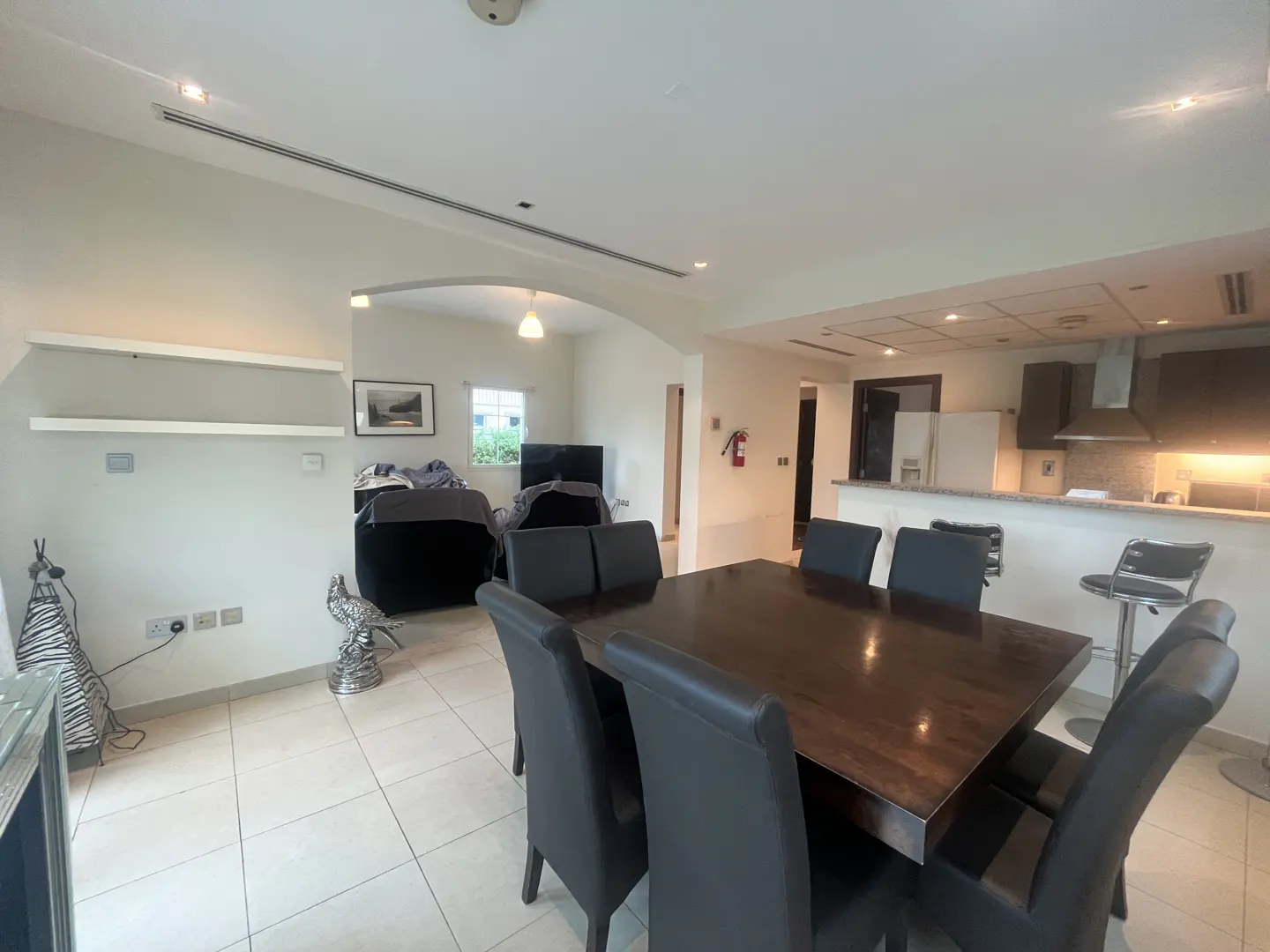 A bright, open-plan living and dining area with a dark wood table and black leather chairs. The kitchen has a breakfast bar with stools.