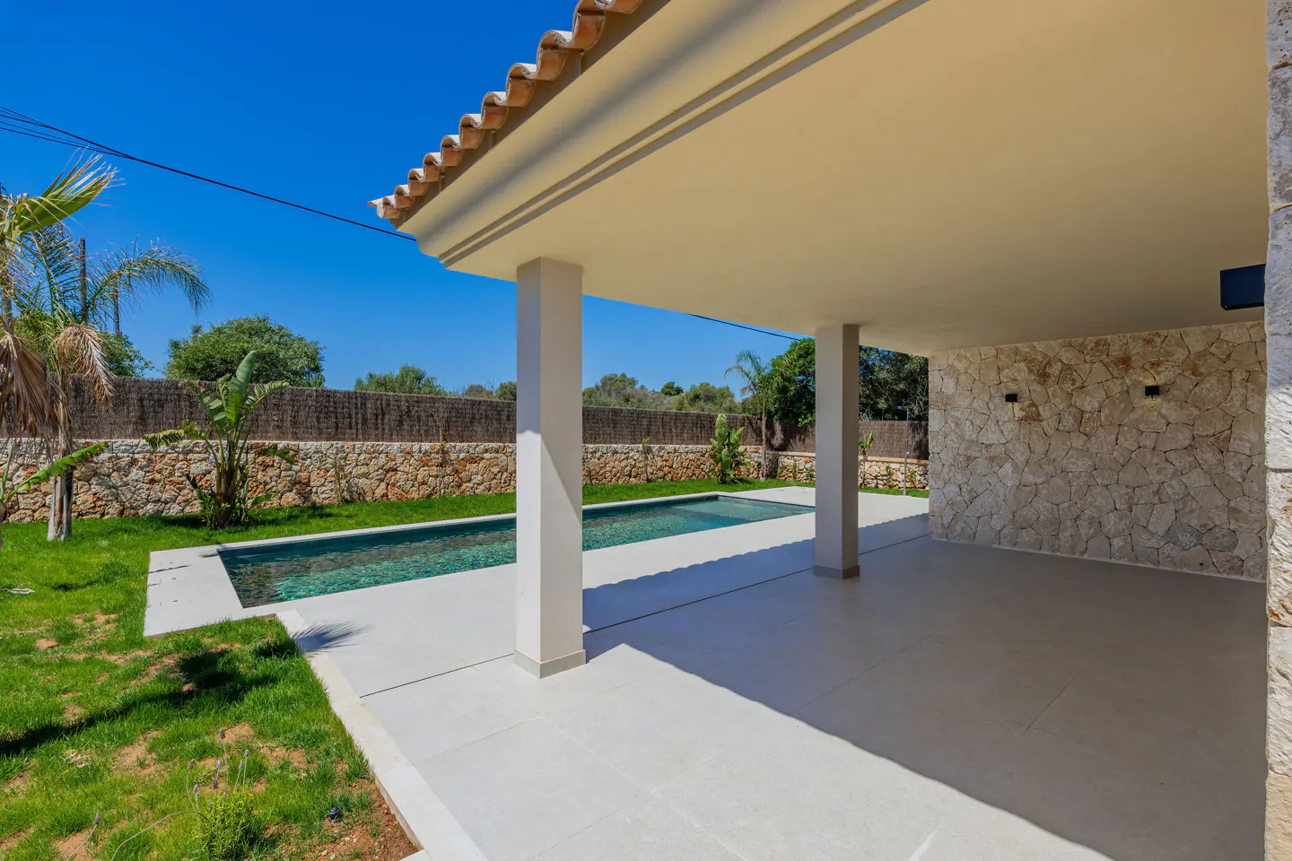 Covered patio with white tile floor, stone accent wall, and view of a pool and green lawn. Blue sky.