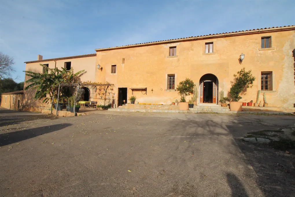 Exterior view of a two-story, peach-colored building with multiple windows and arched doorways, surrounded by greenery and a paved courtyard.