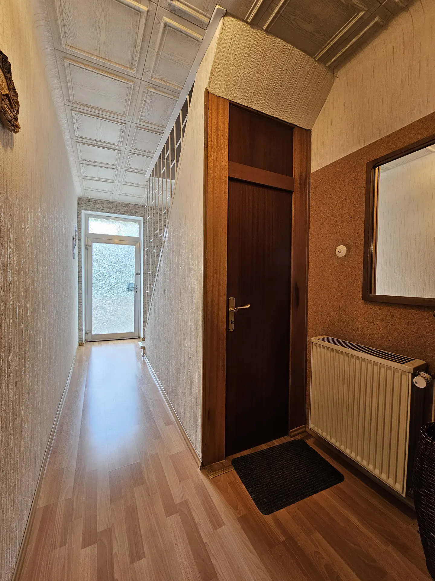 Hallway with wood-look floors, textured walls, and a patterned ceiling. A dark wood door is to the right, with a radiator and mirror. A frosted glass door is at the end of the hall.