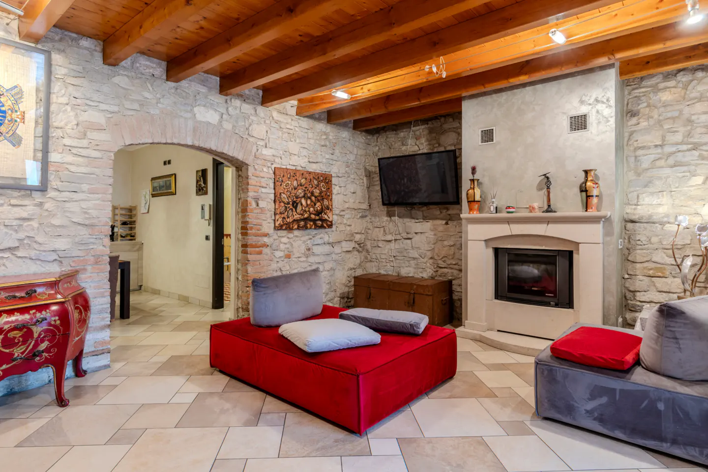 Rustic living room with stone walls, wood beam ceiling, fireplace, red modular seating, and tiled floor.