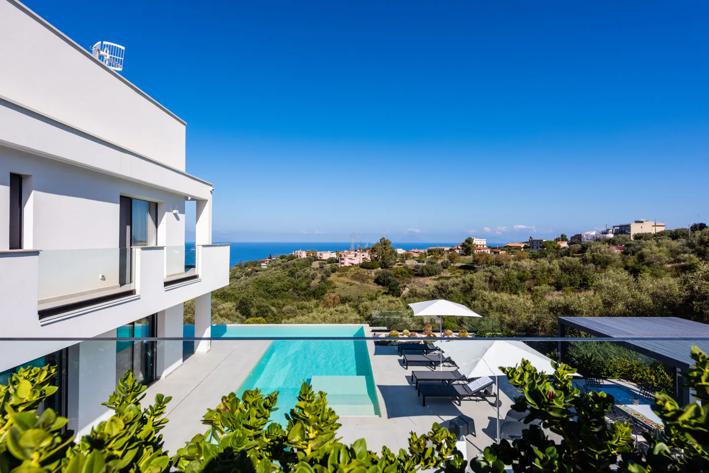 Modern white villa with a pool overlooking the ocean and hillside under a clear blue sky.