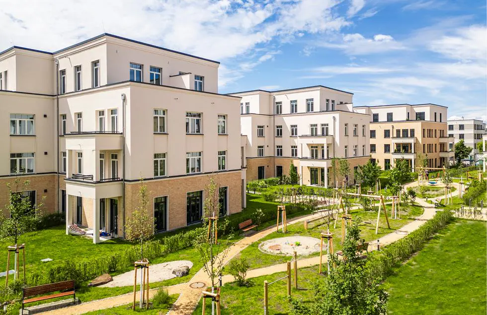 Modern apartment buildings with balconies overlook a green courtyard with walking paths and playground equipment under a blue sky.