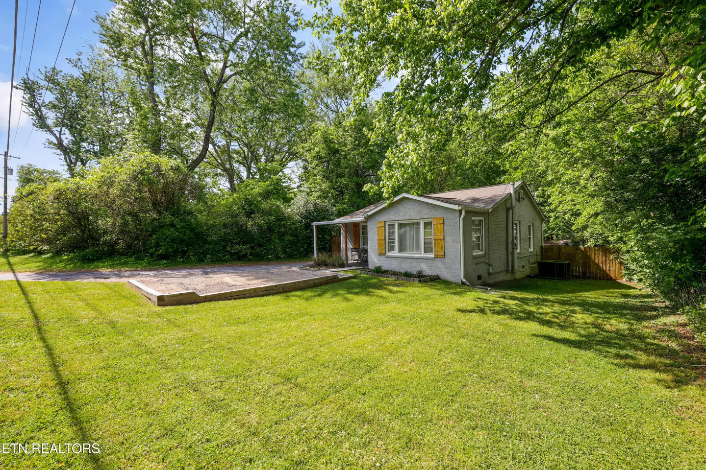 A small, gray house with yellow shutters sits on a large green lawn surrounded by trees.