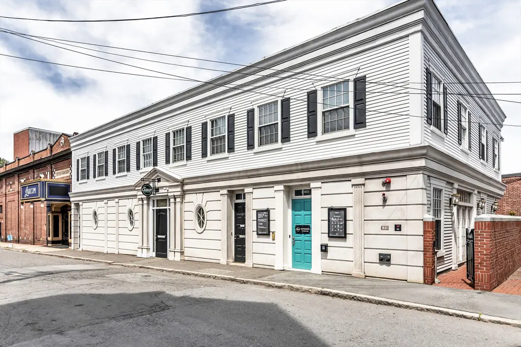 Exterior view of a two-story white building with black shutters and a turquoise door on a city street.