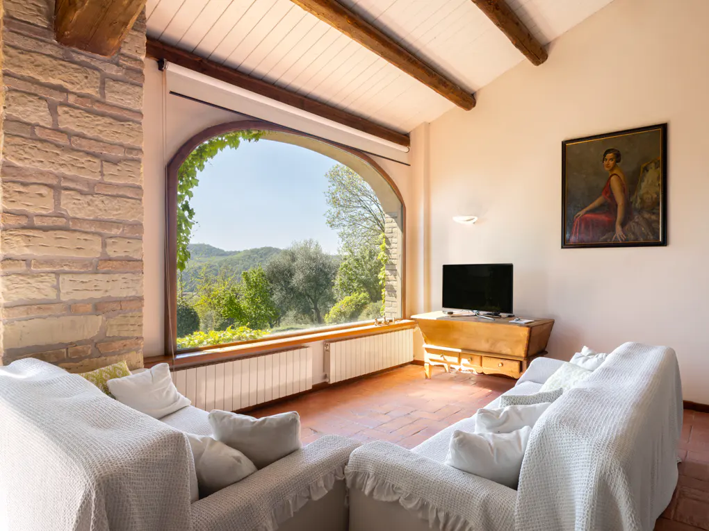 Living room with white sofas, terracotta floor, and large arched window with a view of green hills. A TV sits on a wooden cabinet.