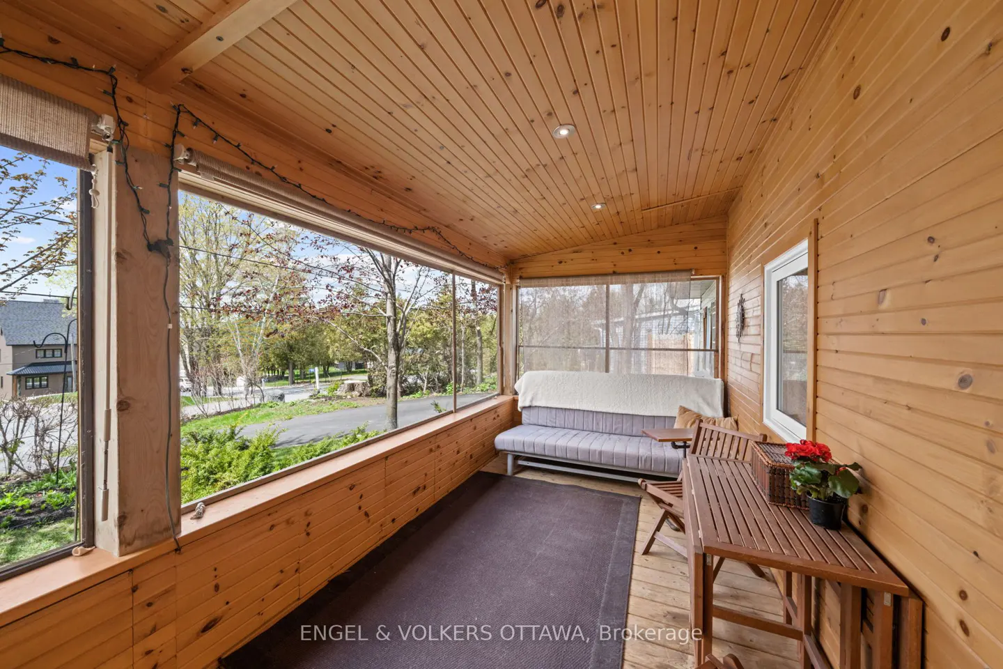 Screened-in porch with wood walls, ceiling, and floor. A gray futon, table, and chairs furnish the space. Outside is a view of trees.