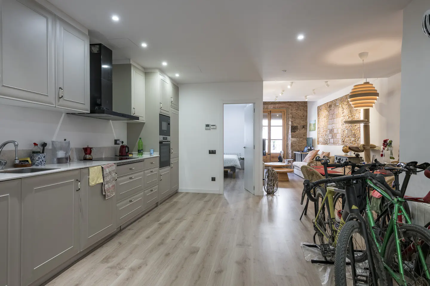 Open-concept apartment with a gray kitchen, wood floors, and bikes parked near a living room with a brick wall.