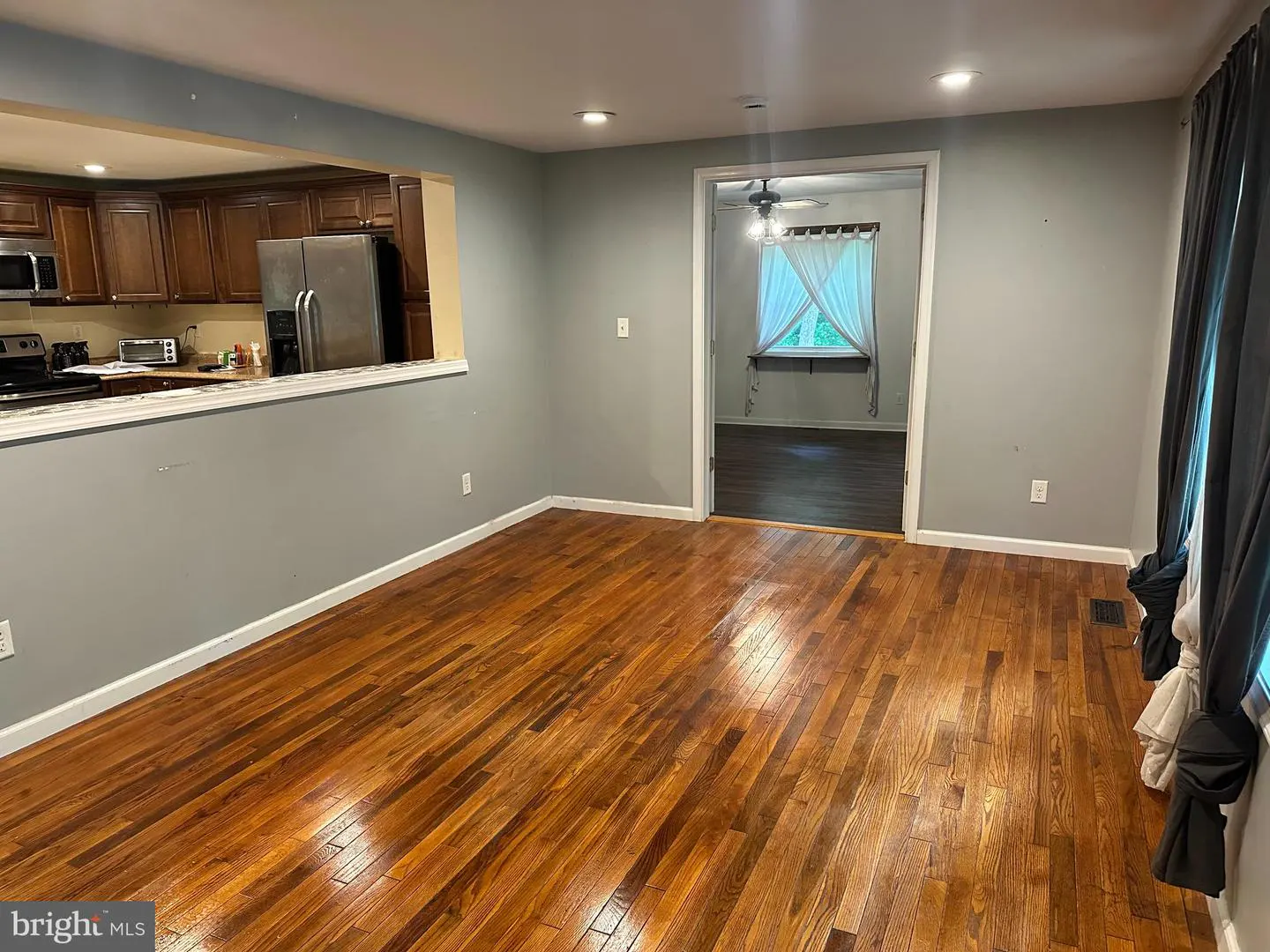 A living room with hardwood floors, gray walls, and a kitchen view through a wall opening. A doorway leads to another room with a window.