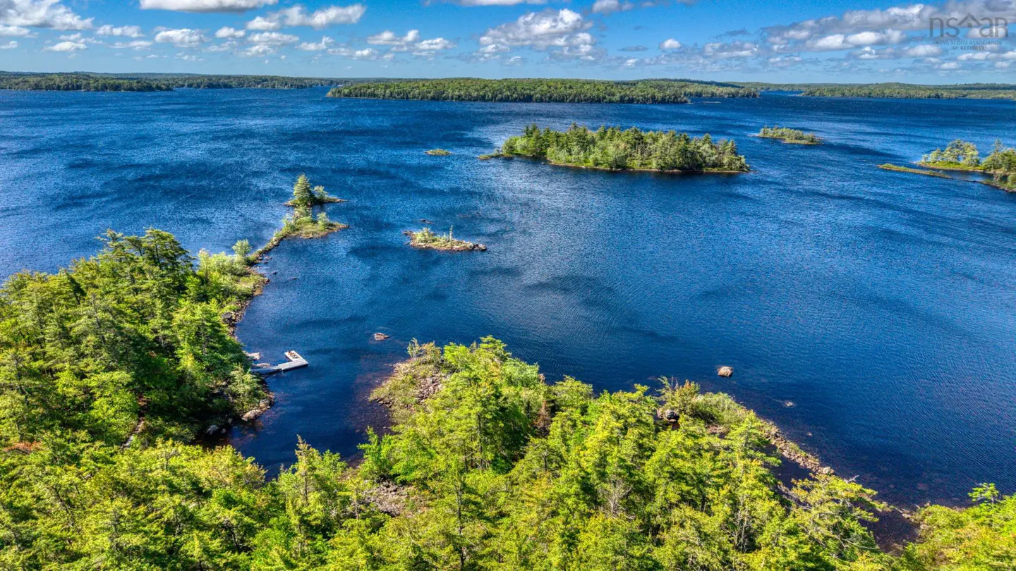 Aerial view of a deep blue lake with forested islands, a small wooden dock, and a bright blue sky with scattered clouds.
