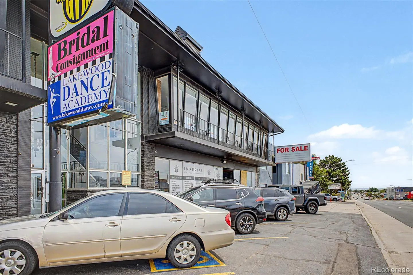 Exterior view of a commercial building with a Bridal Consignment sign, parked cars, and a For Sale sign on a sunny day.