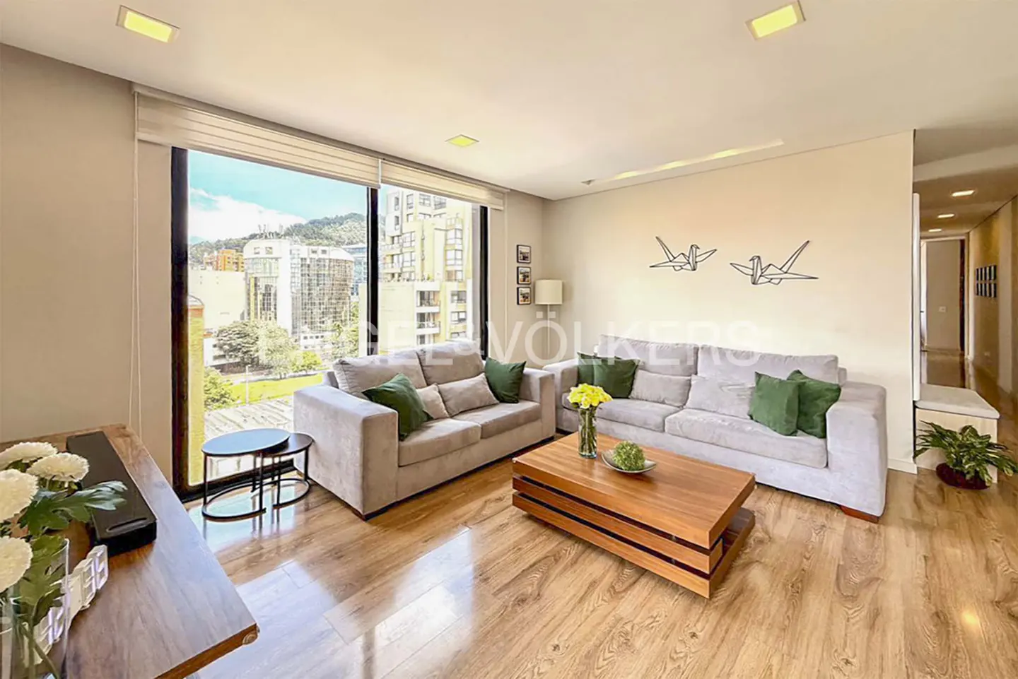 Living room with two gray sofas, green pillows, wood floors, and a city view from a large window.