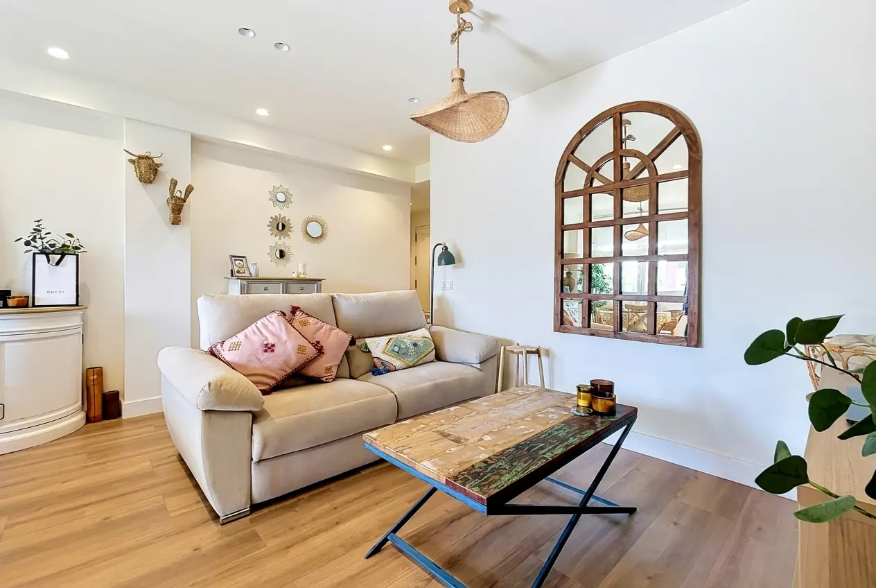 Bright living room with a beige sofa, rustic wooden coffee table, arched wall mirror, and bohemian decor on wood floors.