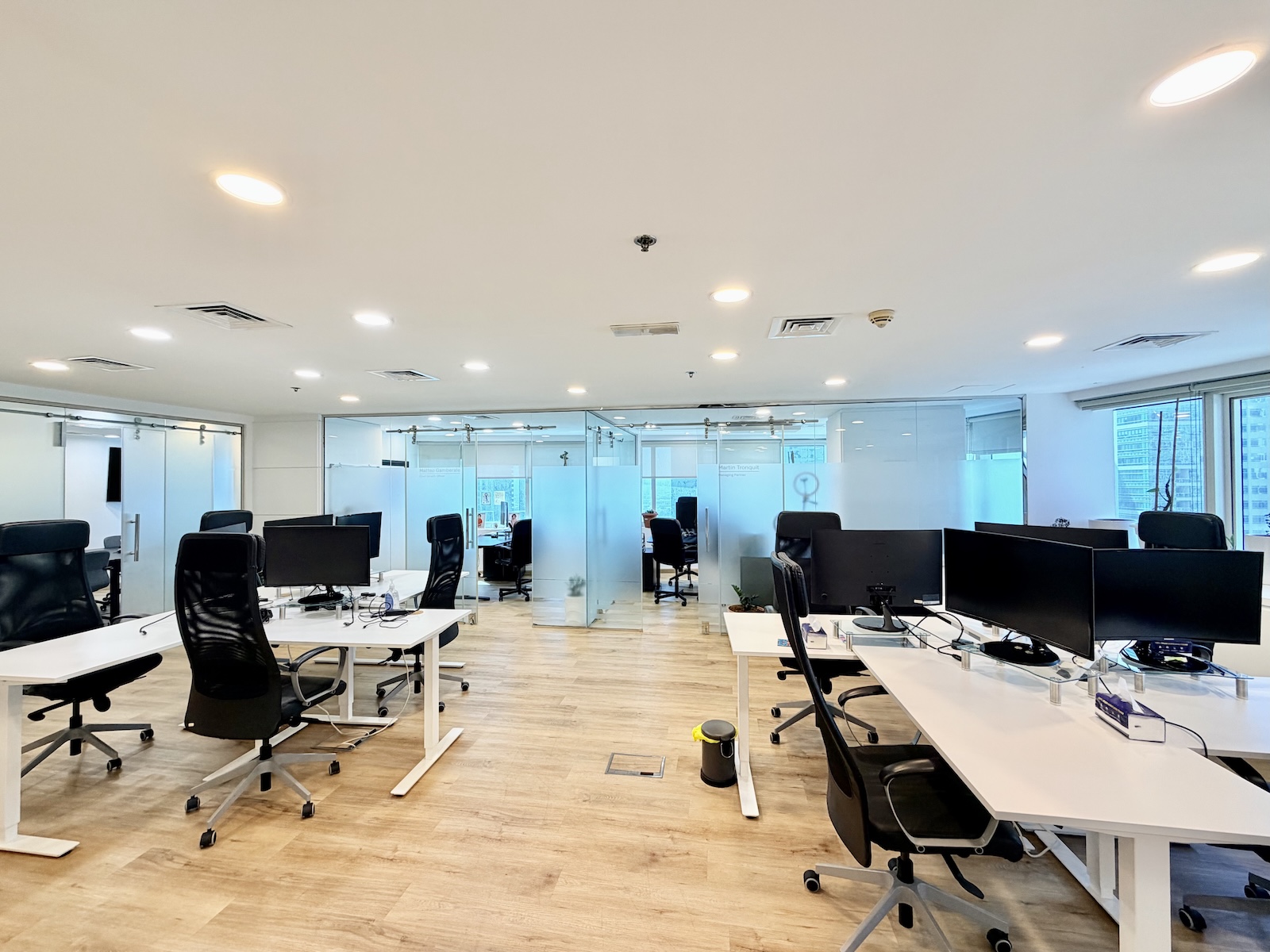 Modern open-plan office with white desks, black ergonomic chairs, computer monitors, and frosted glass partitions.