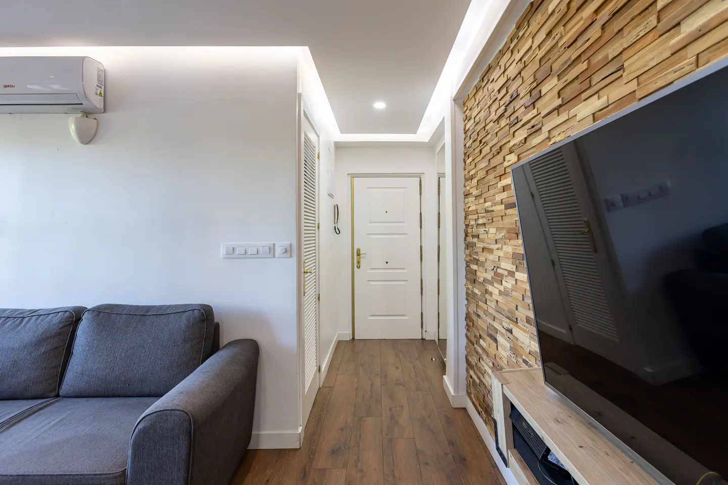 Living room view with a gray sofa, white walls, and a wood-paneled accent wall with a large TV. A hallway leads to a white front door.