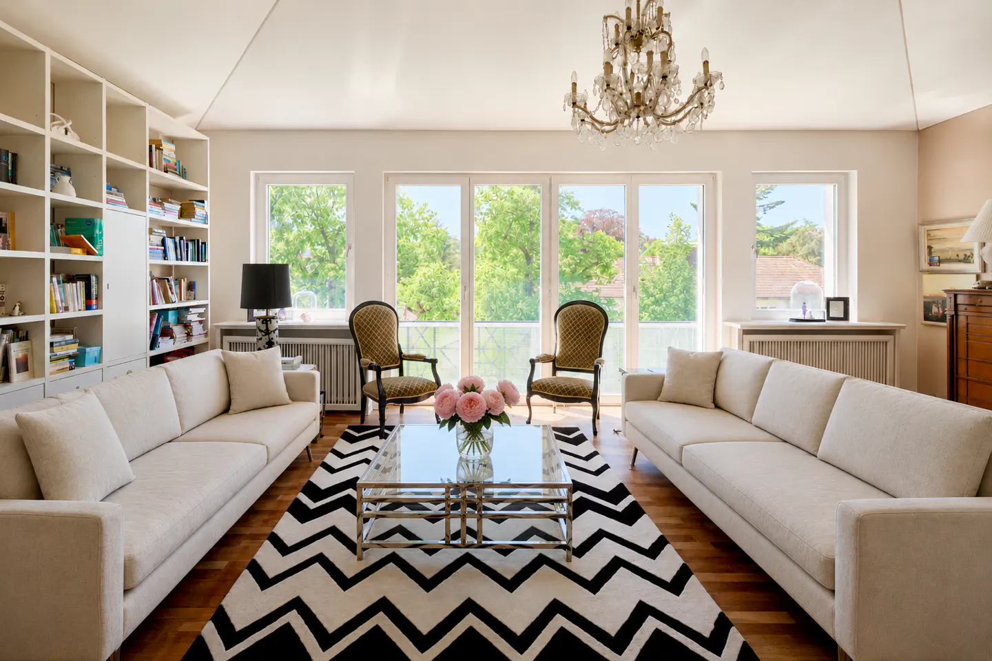 Bright living room with two white sofas, a glass coffee table, a chevron rug, and a crystal chandelier.