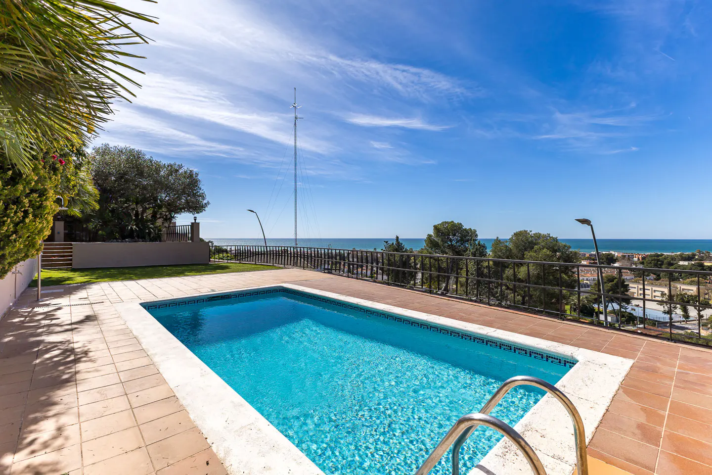 Outdoor pool with turquoise water, surrounded by a tiled patio and a black metal fence overlooking the ocean under a blue sky.