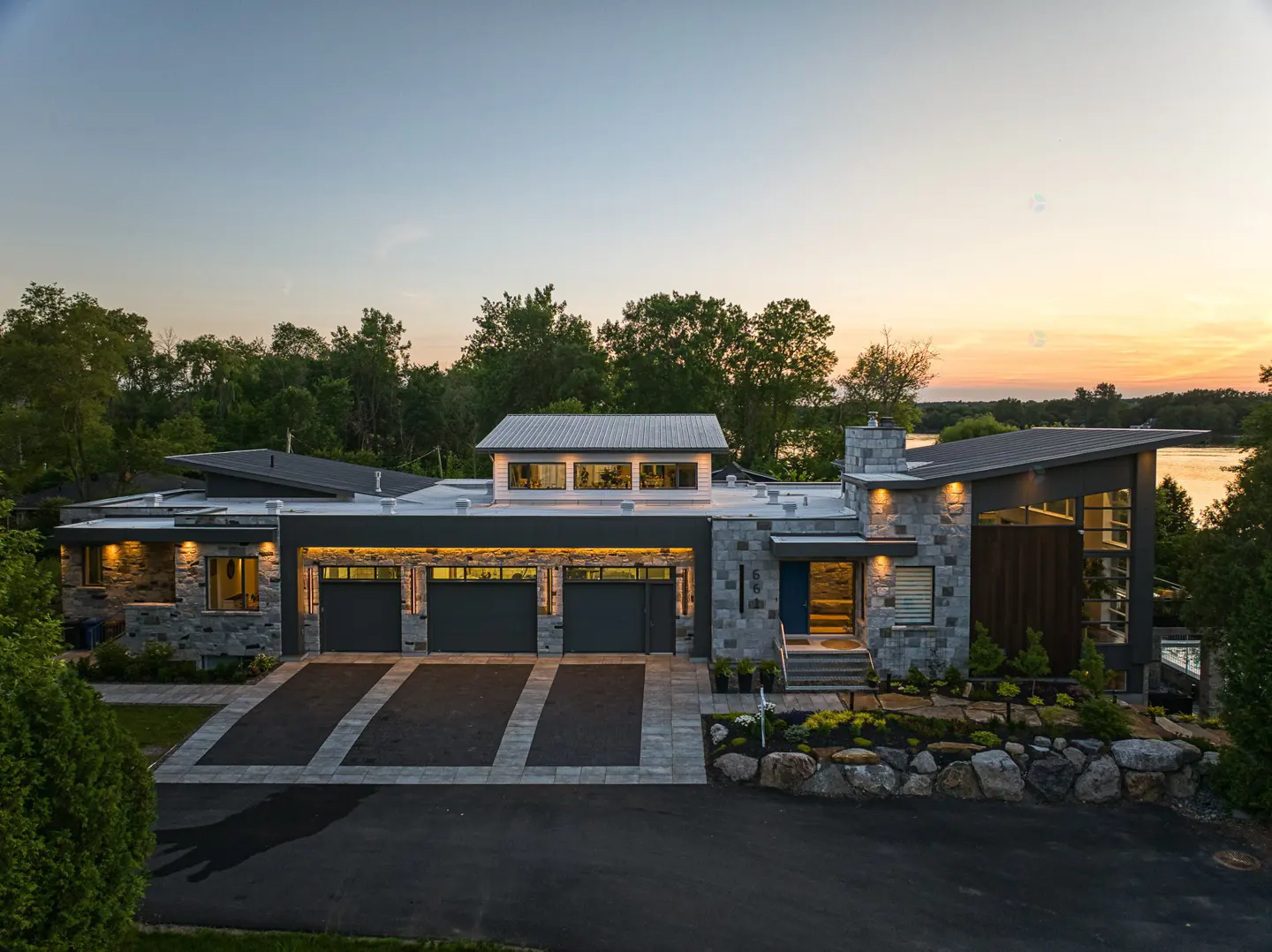 Modern stone house with three-car garage at dusk. The house has a flat roof and a stone chimney. The driveway is paved with black asphalt.