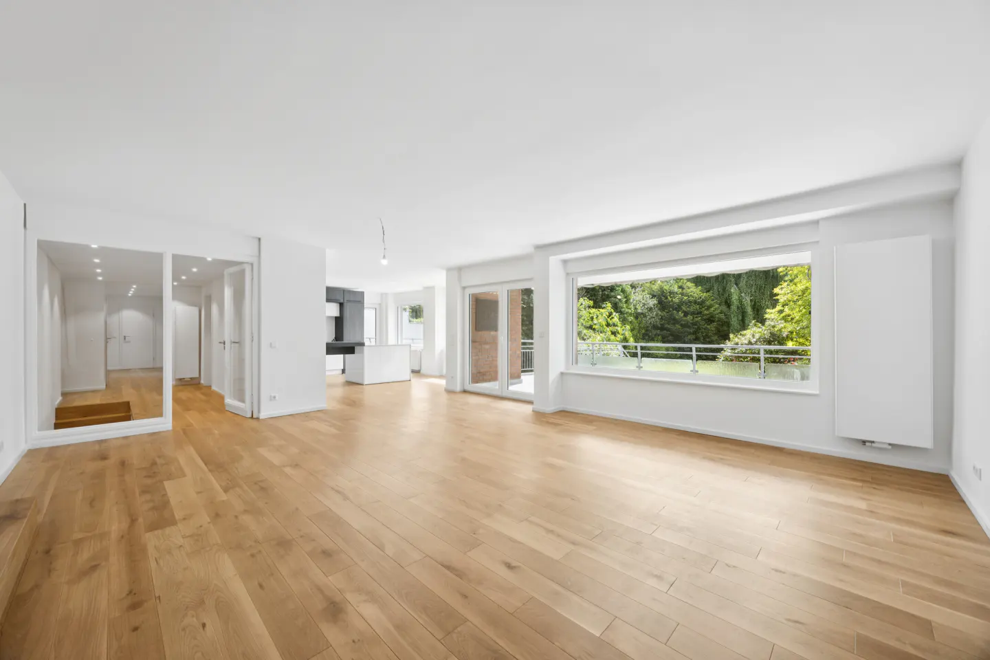 Bright, empty apartment with hardwood floors, white walls, and a large window overlooking a green garden.