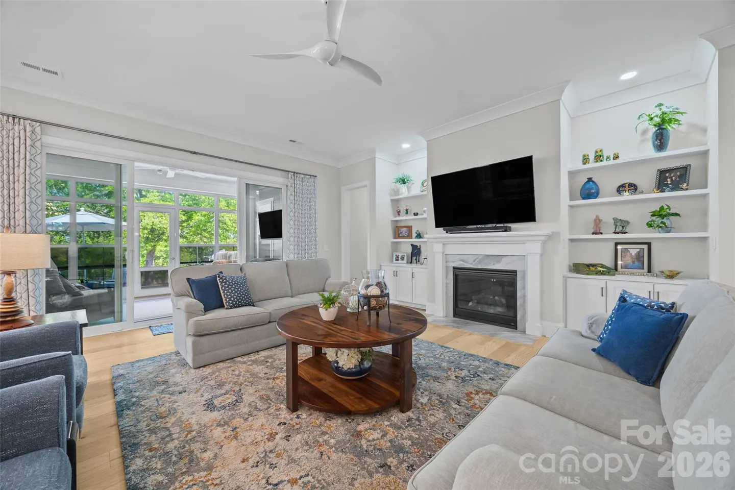 Bright living room with light gray sofas, a round wooden coffee table, fireplace, TV, and built-in shelves on wood floors.