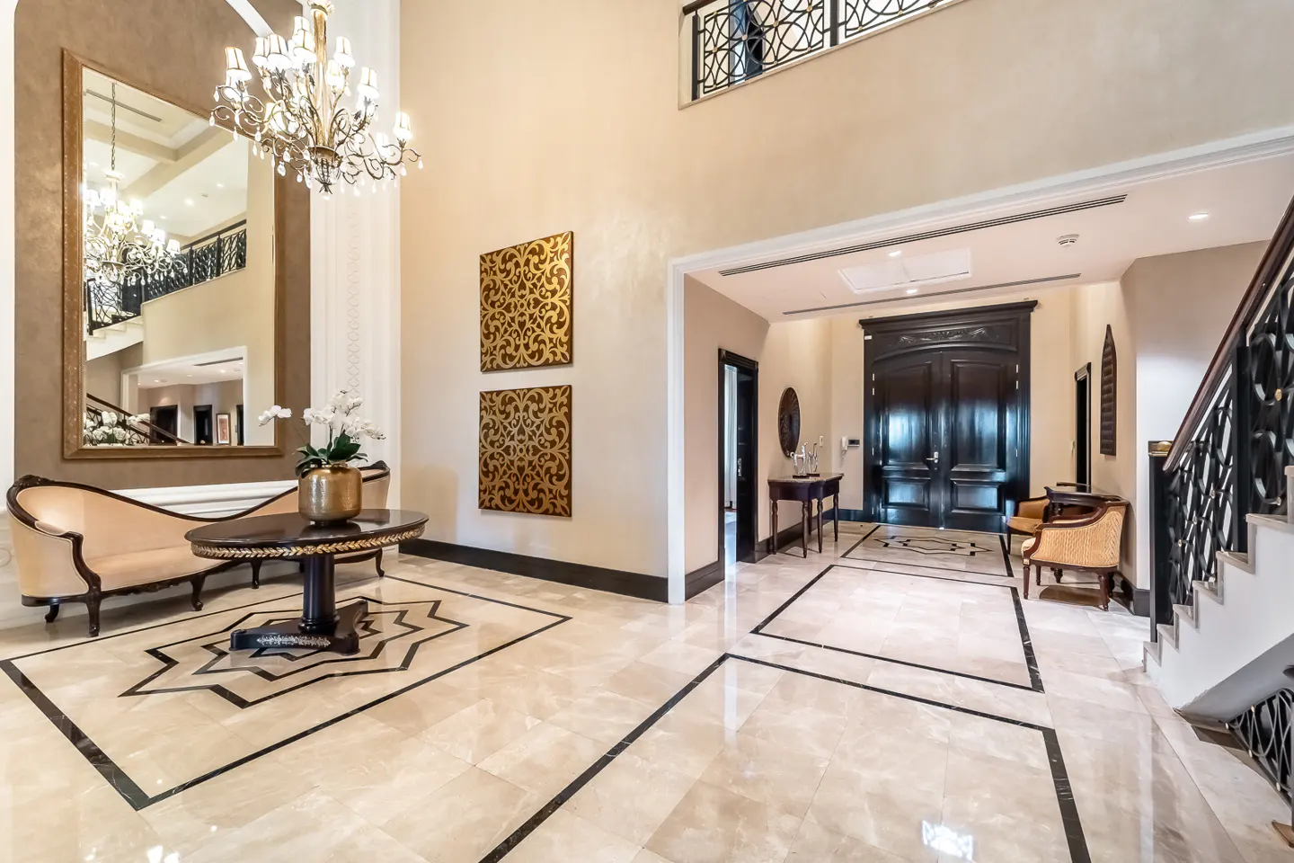 Opulent foyer with marble floors, a crystal chandelier, and a large mirror reflecting a staircase. A beige chaise lounge sits near a round table with flowers. Two dark wood doors are visible.