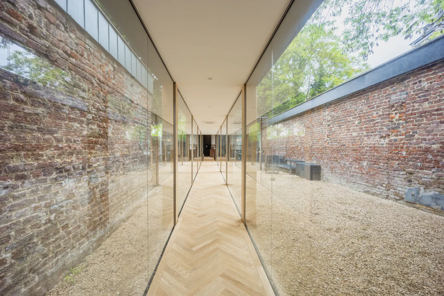 Hallway with herringbone wood floor, glass walls, and brick walls on either side. Gravel outside the glass.