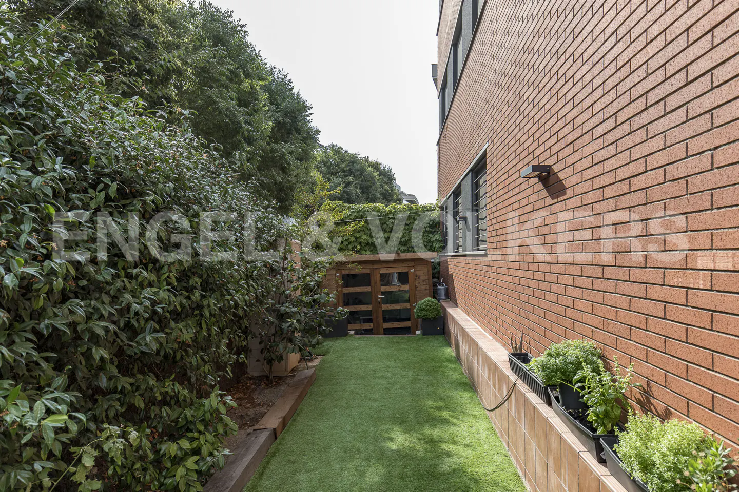 A narrow backyard with artificial grass, a wooden shed, and a brick building. Green bushes line the left side.