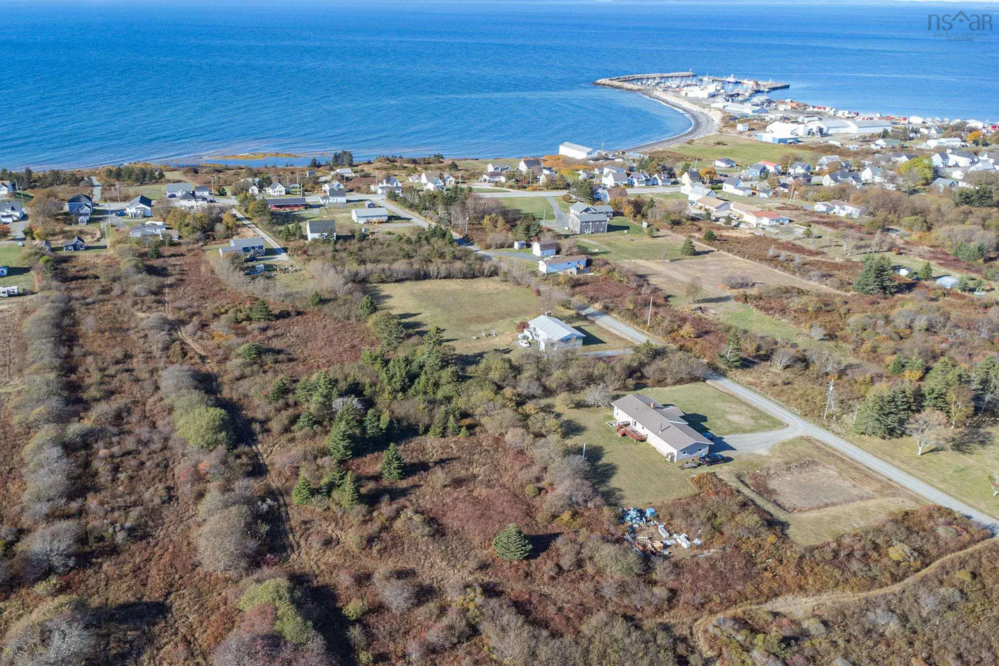 Aerial view of land for sale near the ocean, with houses, trees, and a harbor in the background. The ocean is blue.