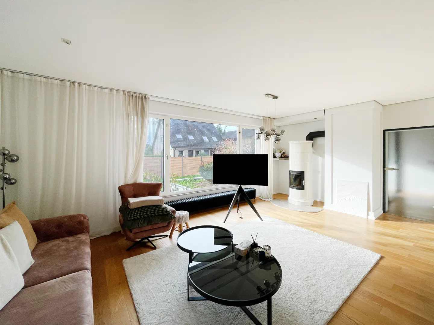 Bright living room with wood floors, white rug, and a black TV on a tripod stand. A fireplace and a brown sofa add warmth.