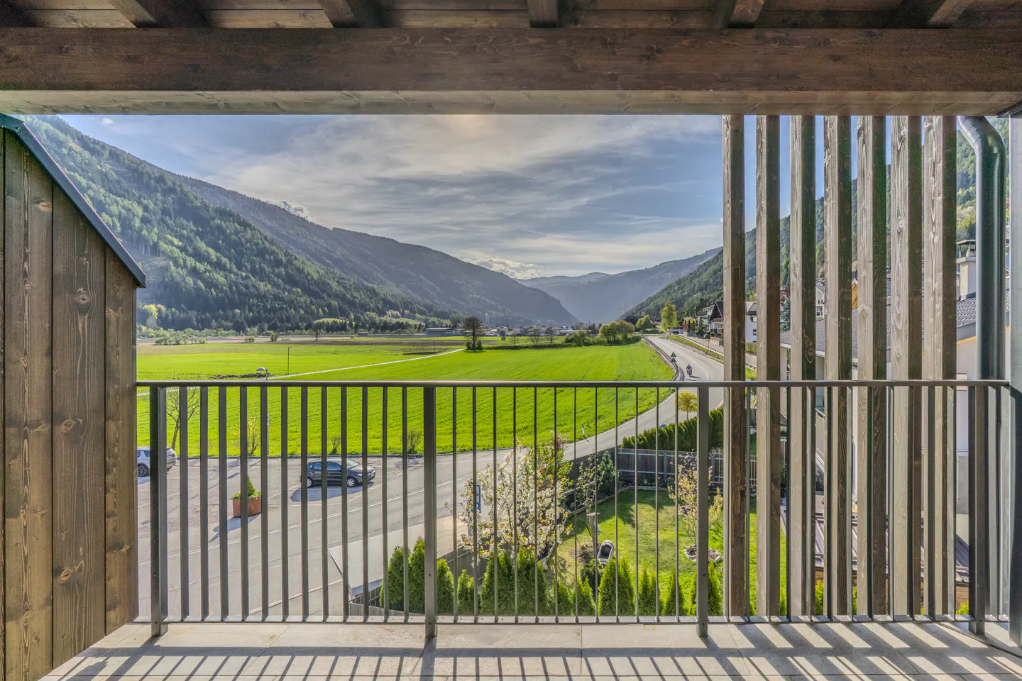 View from a balcony with metal railings overlooking a green valley and mountains under a blue sky.