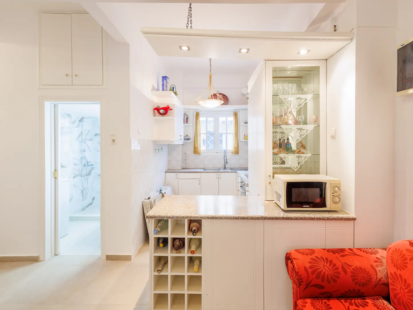 Bright white kitchen with a granite-topped island, wine rack, glass display cabinet, and a red floral sofa in the foreground.