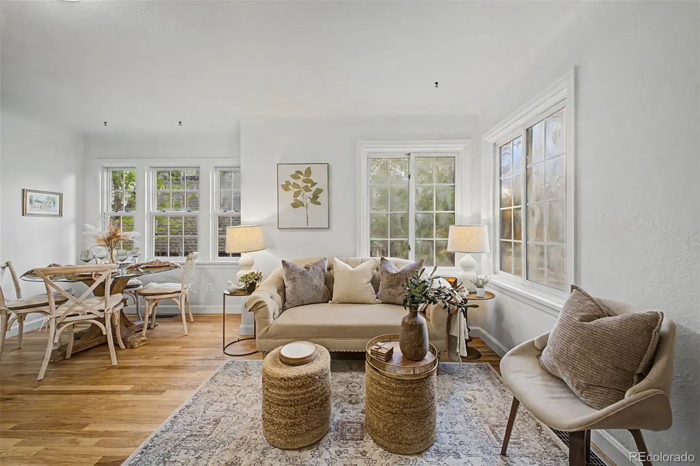 Bright living room with hardwood floors, white walls, and large windows. A beige sofa with pillows sits on a patterned rug, with woven stools as tables. A dining table is visible.