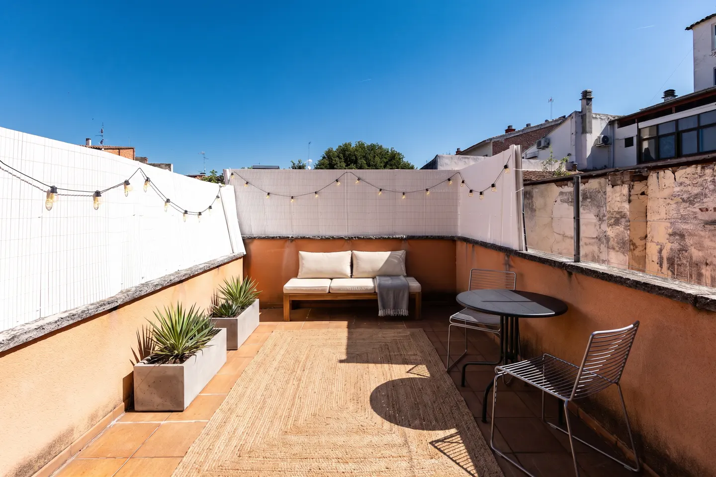 Outdoor patio with a tan rug, a wooden couch with white cushions, a table with two chairs, and string lights.
