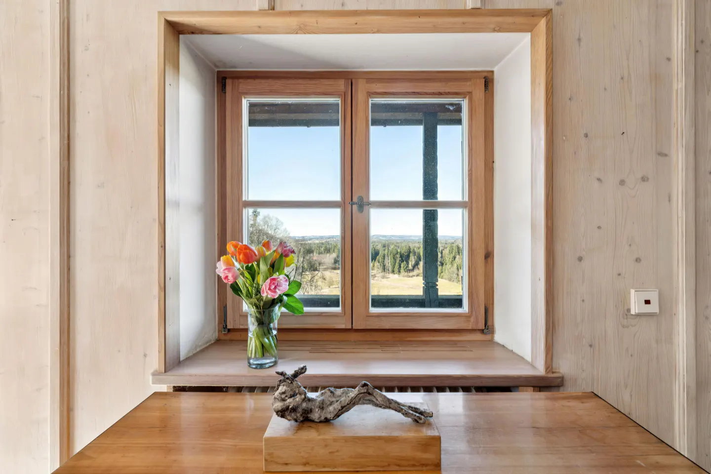 Interior view of a wood-paneled room with a window, flowers, and sculpture. Outside the window is a view of trees and a blue sky.