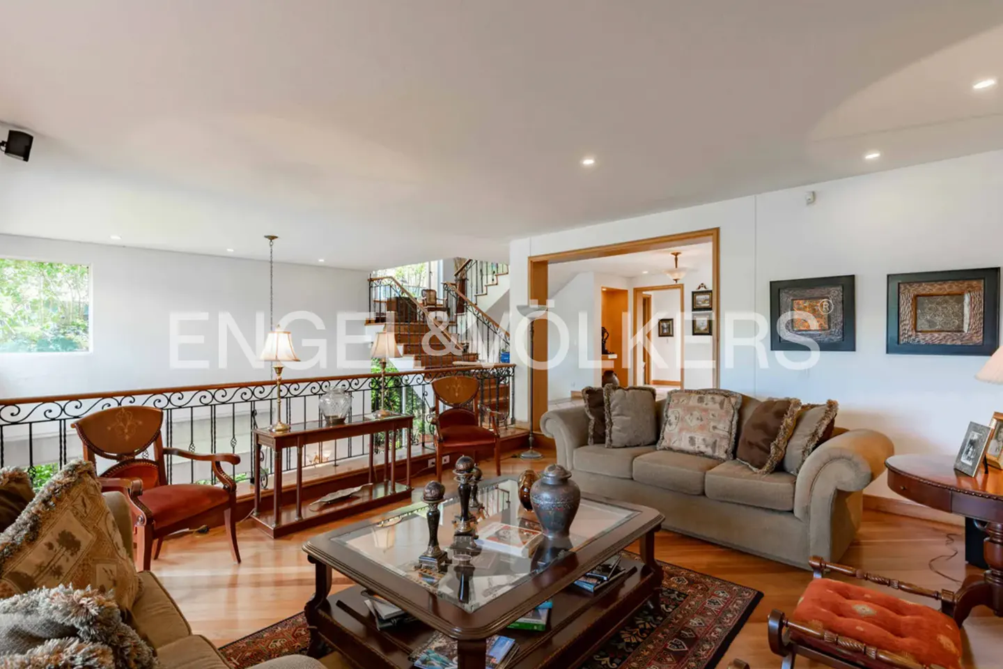 A living room with a brown sofa, a glass coffee table, and a staircase in the background. The walls are white, and the floor is wooden.