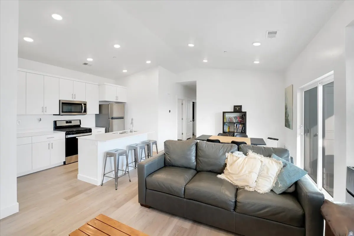 Bright, open-concept living space with white kitchen cabinets, stainless steel appliances, and a gray leather sofa with throw pillows.