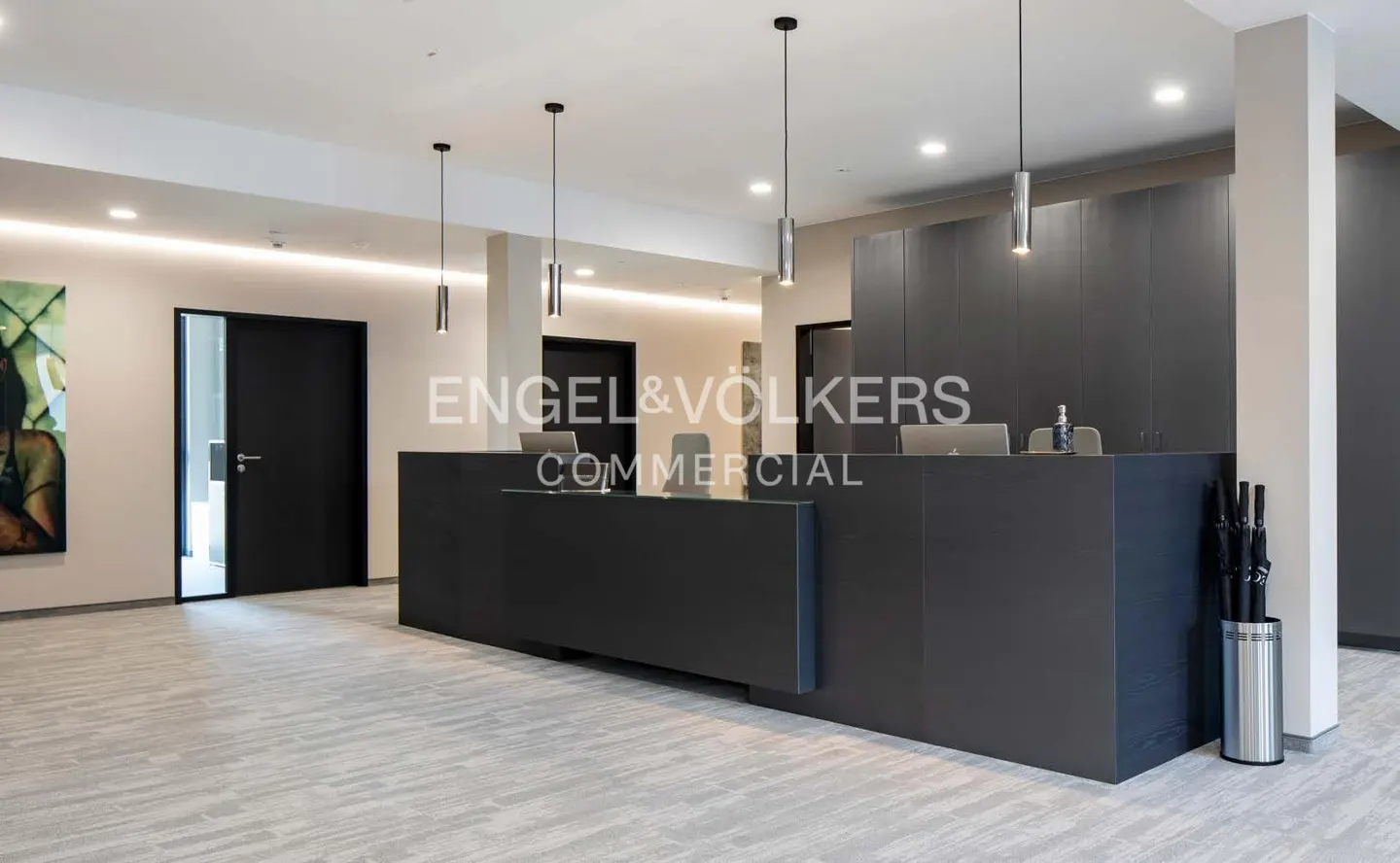 Modern office lobby with a dark gray reception desk, pendant lights, and gray tiled floor. Engel & Volkers logo visible.
