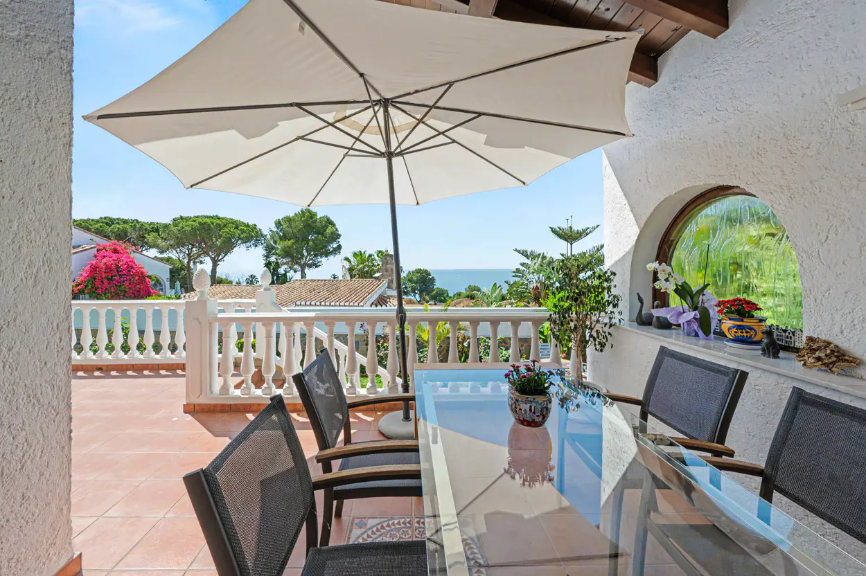 Outdoor patio with a glass table, chairs, and a white umbrella. A white railing overlooks trees and the ocean. A window is on the right.