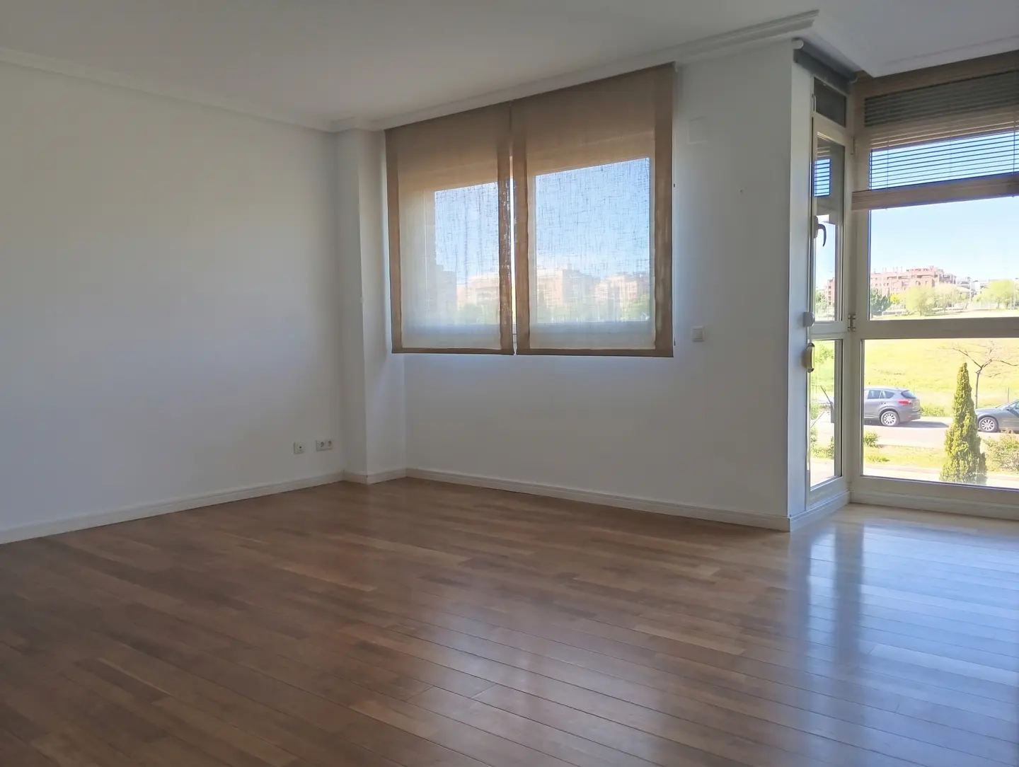 Empty room with white walls, hardwood floors, and large windows with tan shades overlooking a street and green space.