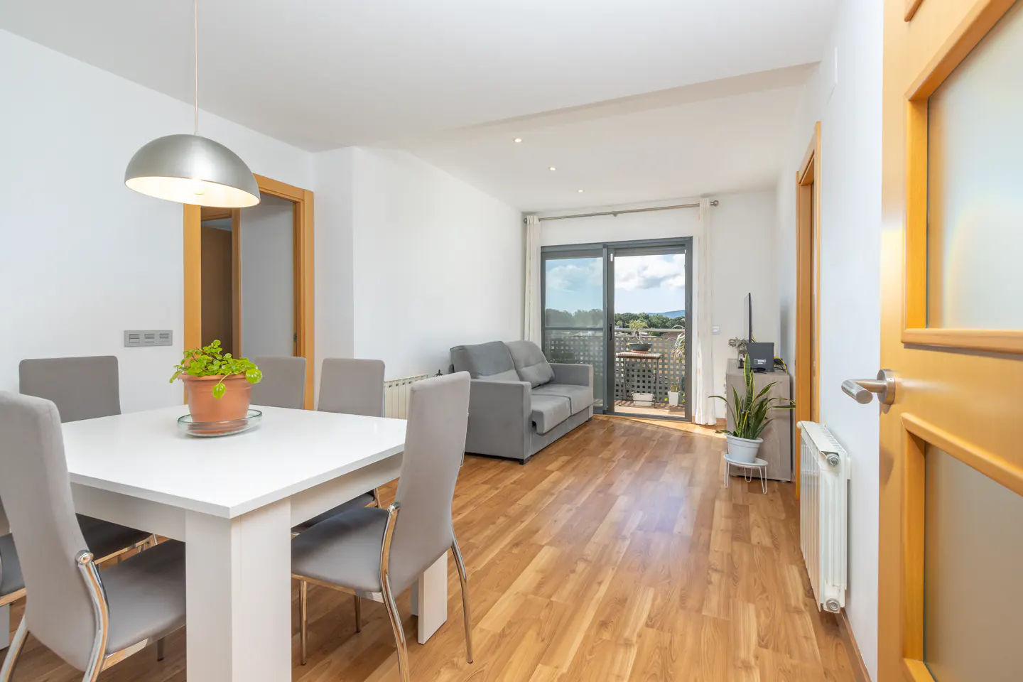 Bright apartment interior with a white table, gray chairs, a gray sofa, and a balcony with a view.